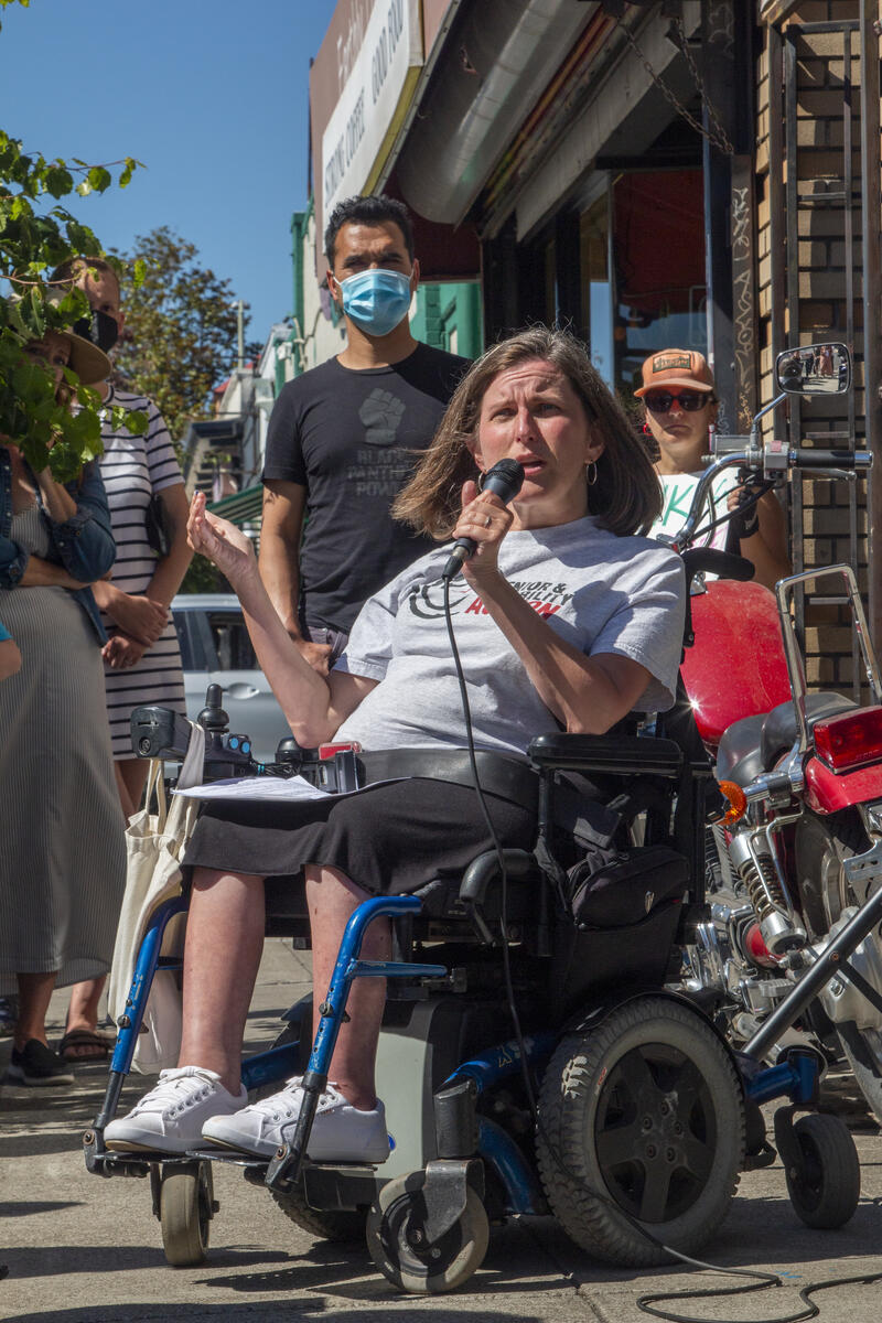 Jessica in a blue power wheelchair with brown hair, holding a microphone in one hand as she speaks.