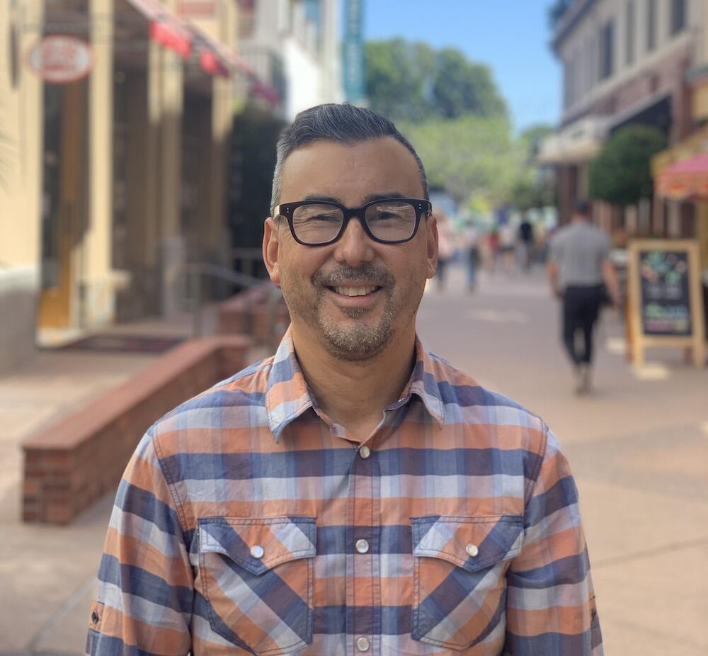 Adam Fukushima smiling in a plaid shirt, standing in a pedestrian walkway in downtown San Luis Obispo