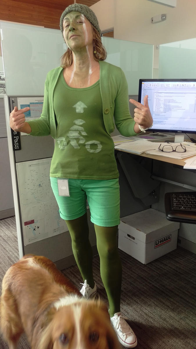A woman dressed up as a green painted bike lane smirks stands in her office.