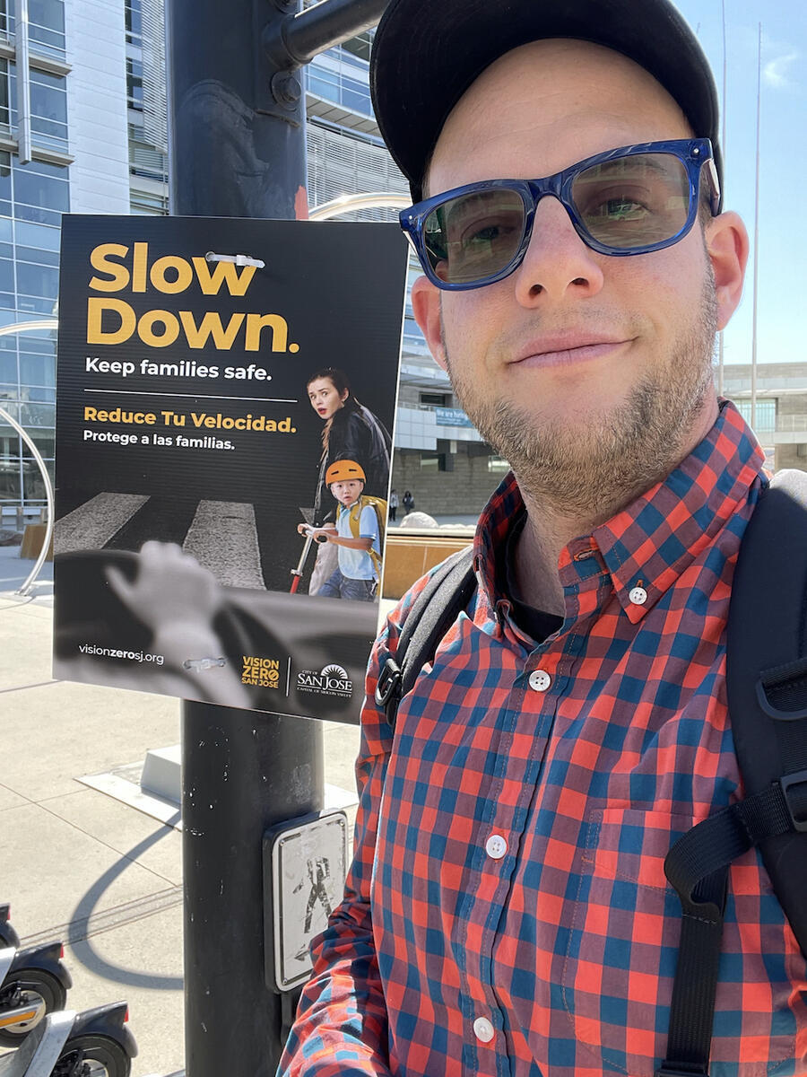 Jesse wearing blue rimmed glasses, a red striped shirt and a hat, smiling next to a sign from the Slow Down campaign
