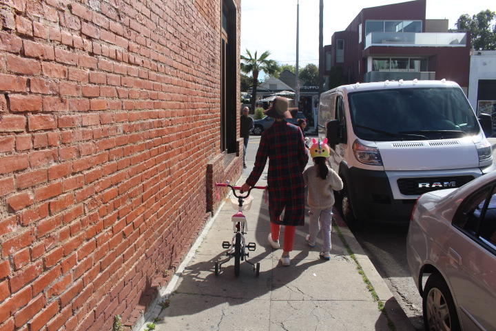 A woman and child stroll on a sidewalk together holding a child's bicycle.
