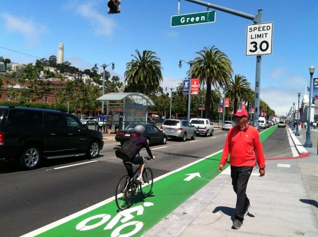 A cyclist bikes in a colored bike lane, while a pedestrian passes. At left, a group of transit users wait for a trolley to arrive.