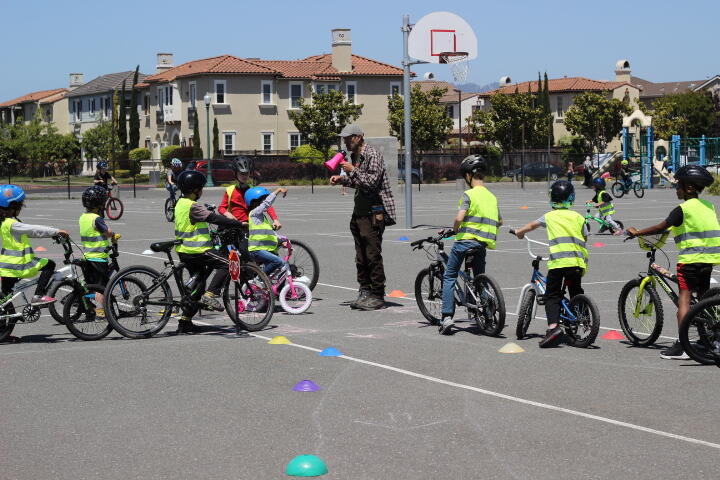 Children in safety vests on bikes surround an instructor with a pink megaphone during a bike rodeo. 