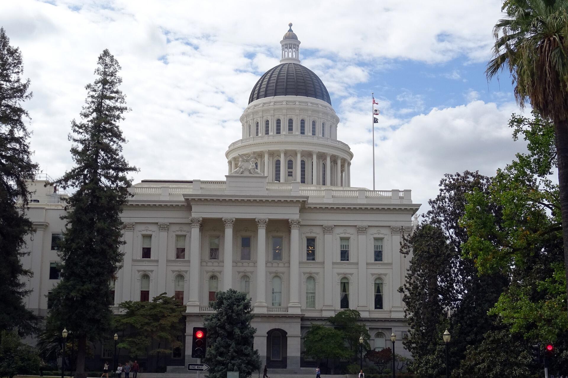 Photo of the front of the State Capitol in Sacramento, it's a white building with columns and a large dome in the middle of the roof with pine trees on either side of the building.