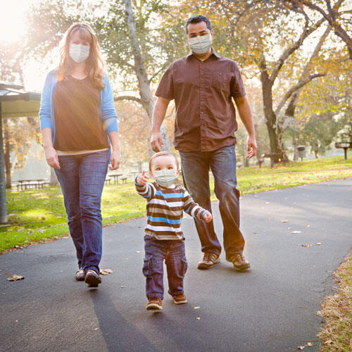 Two parents and a child walk in a park, all are wearing masks. The sun is shining from behind them. 