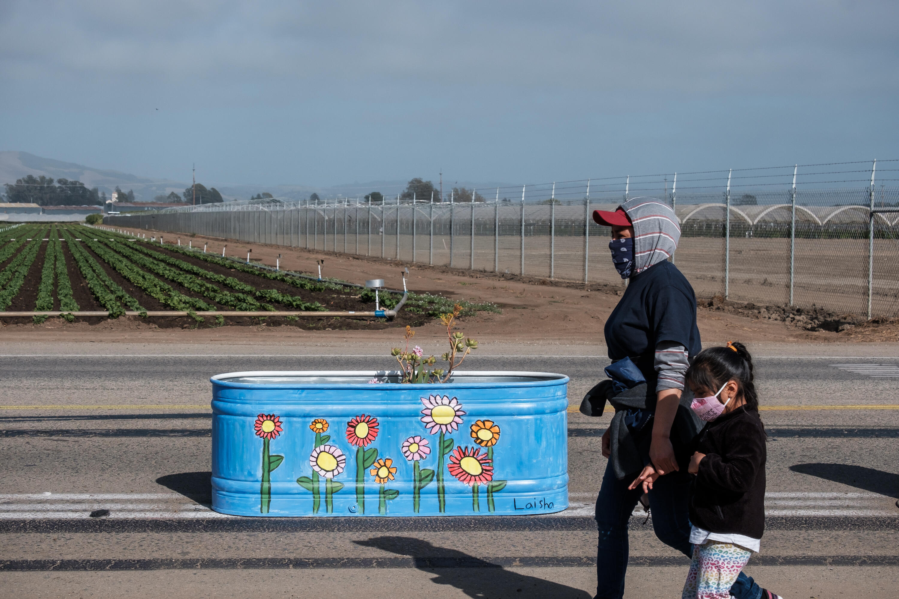 An adult and a kid walk along the Green Valley popup, a painted planter with flowers is behind them and farms are in the background.