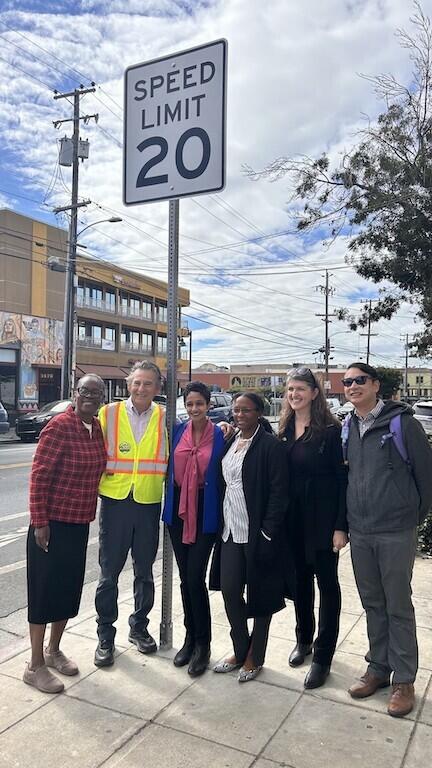 Six people are standing and smiling for a photo by a new street sign that reads "Speed Limit 20 MPH."