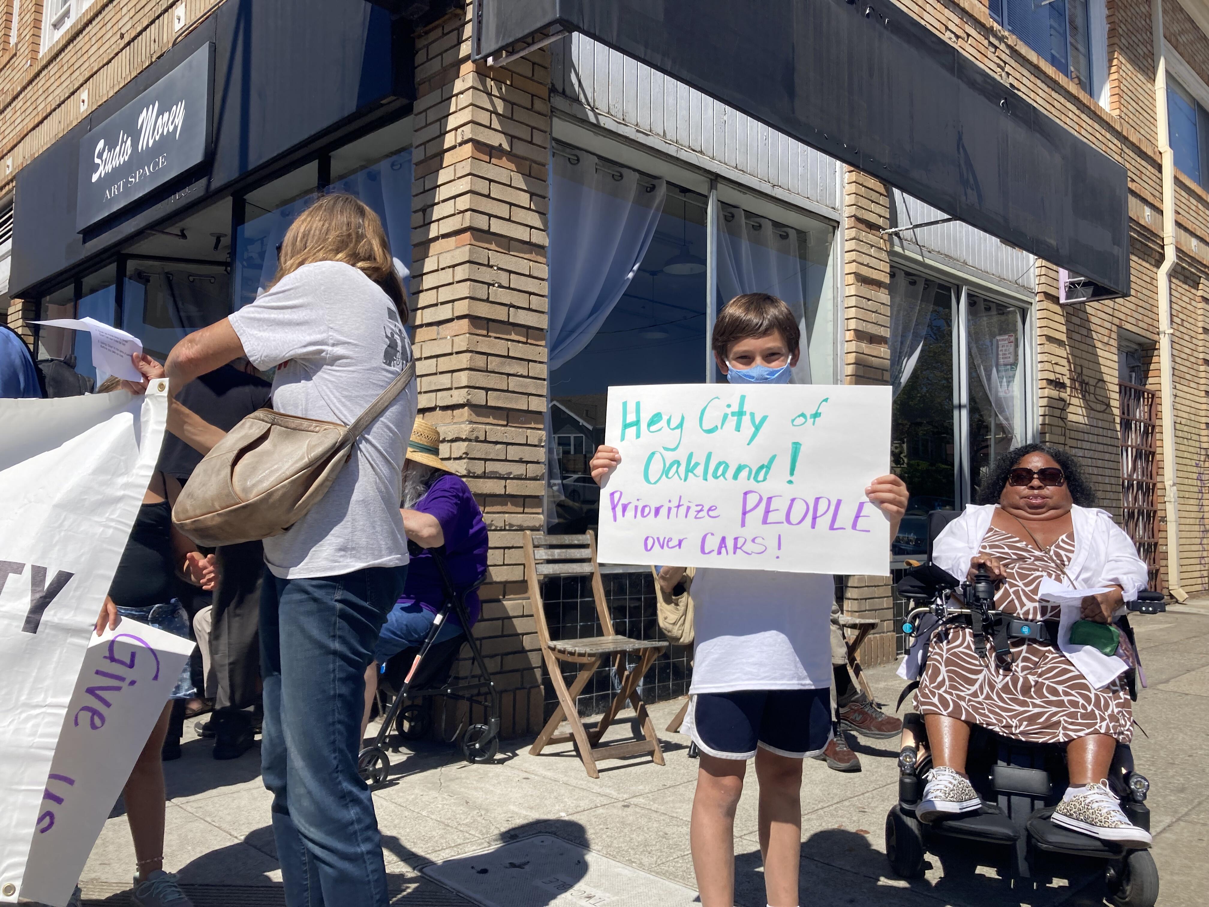 A child holds a poster that reads "Hey City of Oakland! Prioritize people over cars!" They have short brown hair, a blue mask, white shirt, and blue shorts.