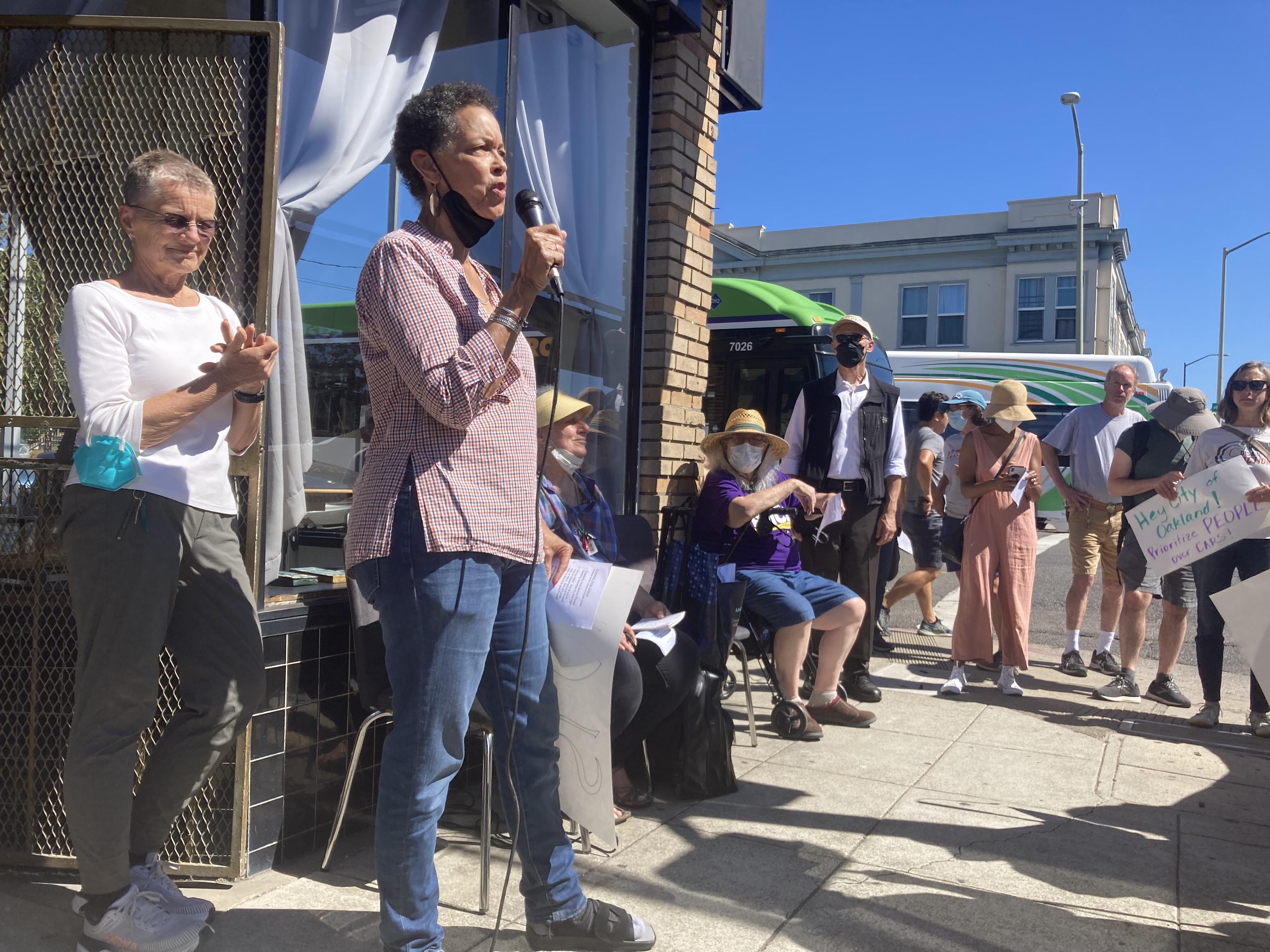 A protestor speaks to the crowd, they have short curly black hair and are wearing a black mask, red and white long sleeve shirt, and blue jeans. They're holding a microphone as they speak to the crowd.