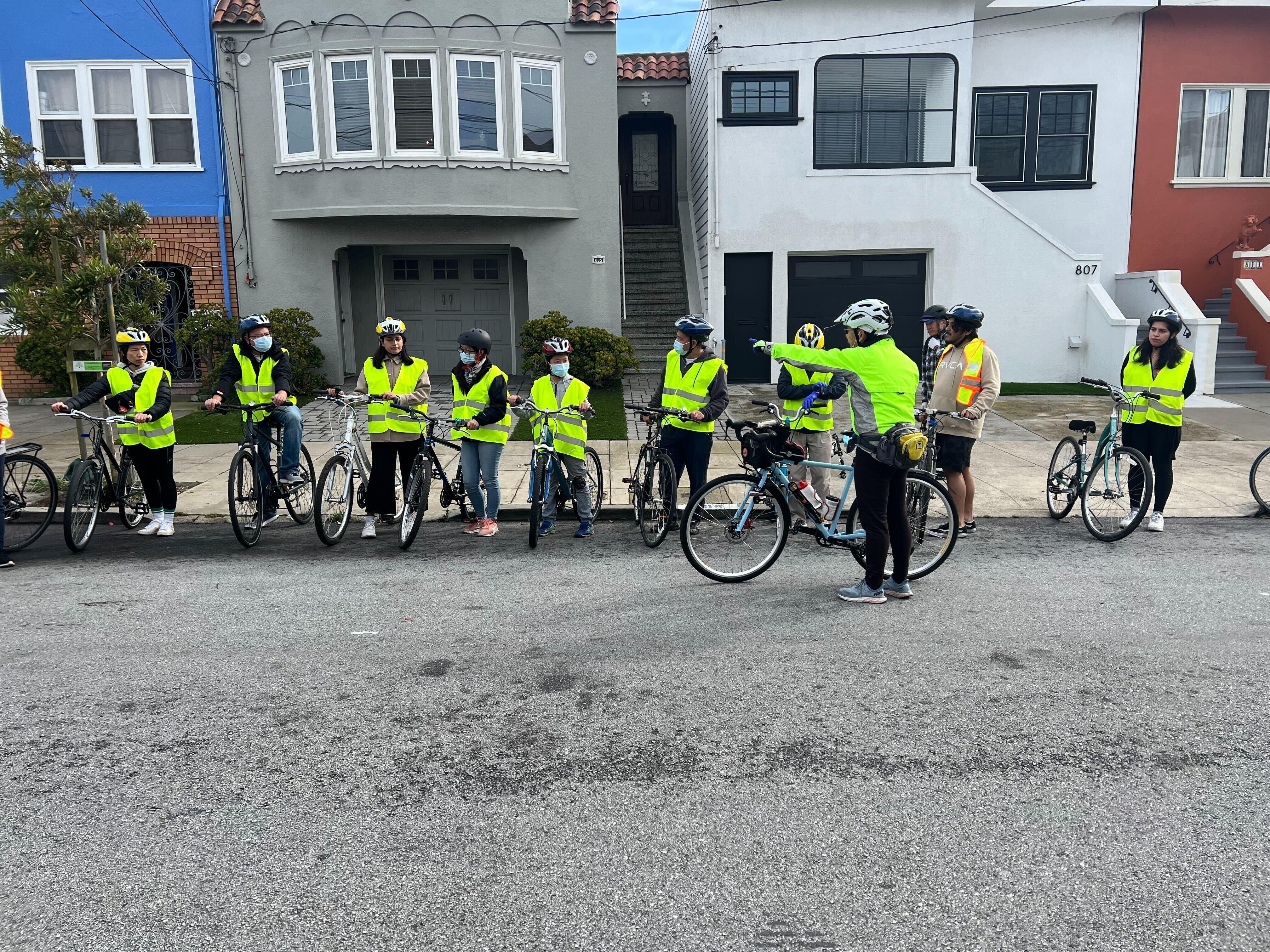 A group of bicyclists are lined up along the curb, all wearing reflective vests.