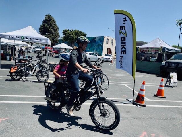 A parent and child ride an electric bike at a farmer's market event for the EZBike Program.