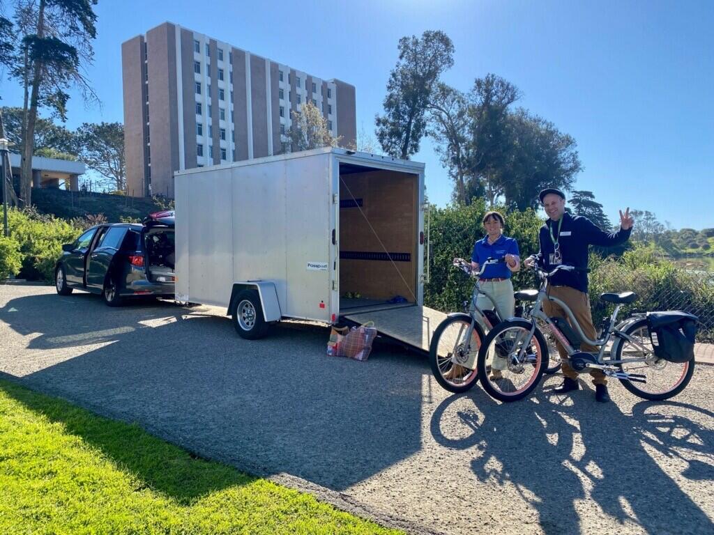 Peter and Eve smiling at the camera, they are unloading electric bikes from a trailer.