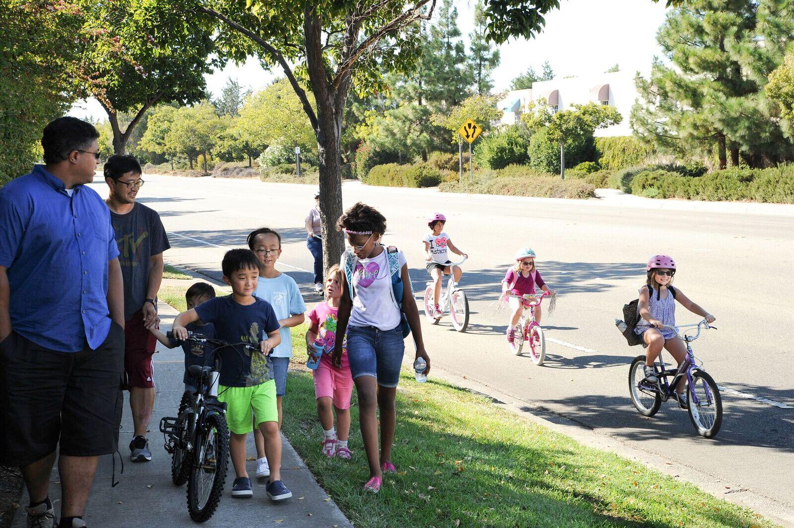 Children and parents walk and bike to school, with some on the sidewalk and others on the road.
