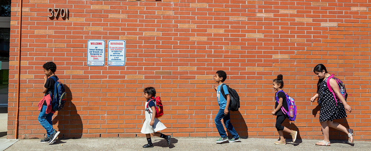 Five children wearing backpacks walk towards their school, with a brick wall behind them. 