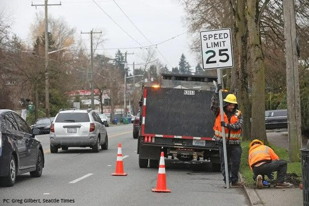 Two construction workers install a 25 MPH speed limit sign on the side of the road.
