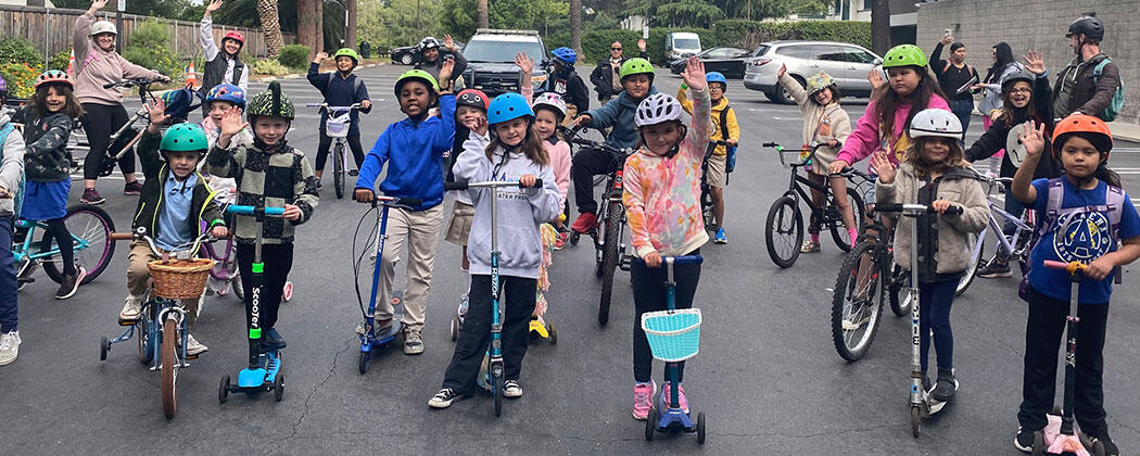 A group photos of children and adults smiling and waving while standing on their bikes and scooters