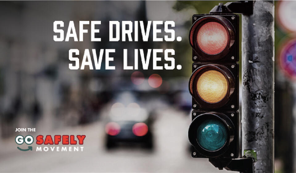 A traffic light at an intersection with the campaign safety message in white text, "Safe Drives. Save Lives."