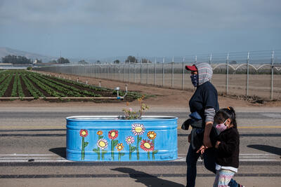 One adult and one kid walk along the Green Valley popup, a painted planter with flowers is behind them and farms are in the background.